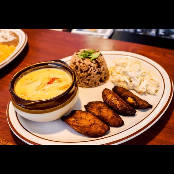 A Mexican meal with yellow soup, rice, coleslaw, and fried plantains on a white plate.