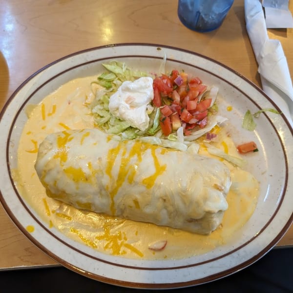 A large, cheese-covered burrito with a side of lettuce, diced tomatoes, and sour cream.